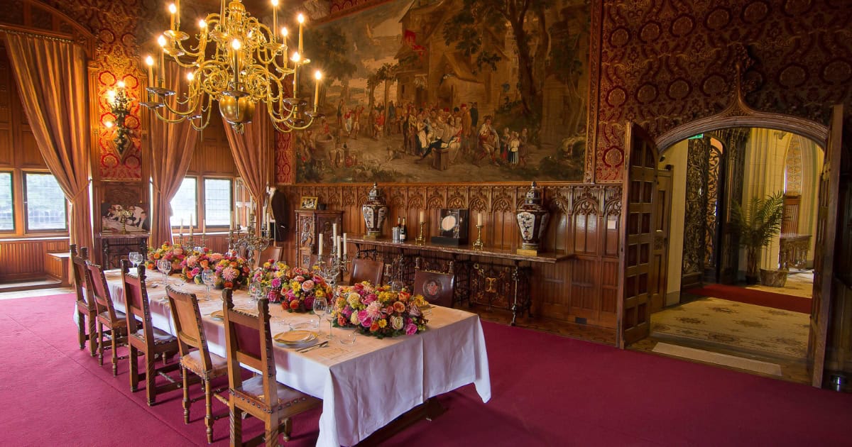A wood-paneled dining room at De Haar Castle with a candlelit chandelier, a table set for a formal dinner, and a large tapestry on the wall.