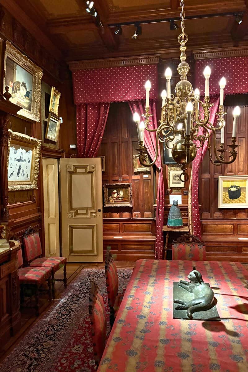 Wood-paneled dining room at the KattenKabinet in Amsterdam, hung with cat paintings under a brass chandelier, a bronze cat on the table.