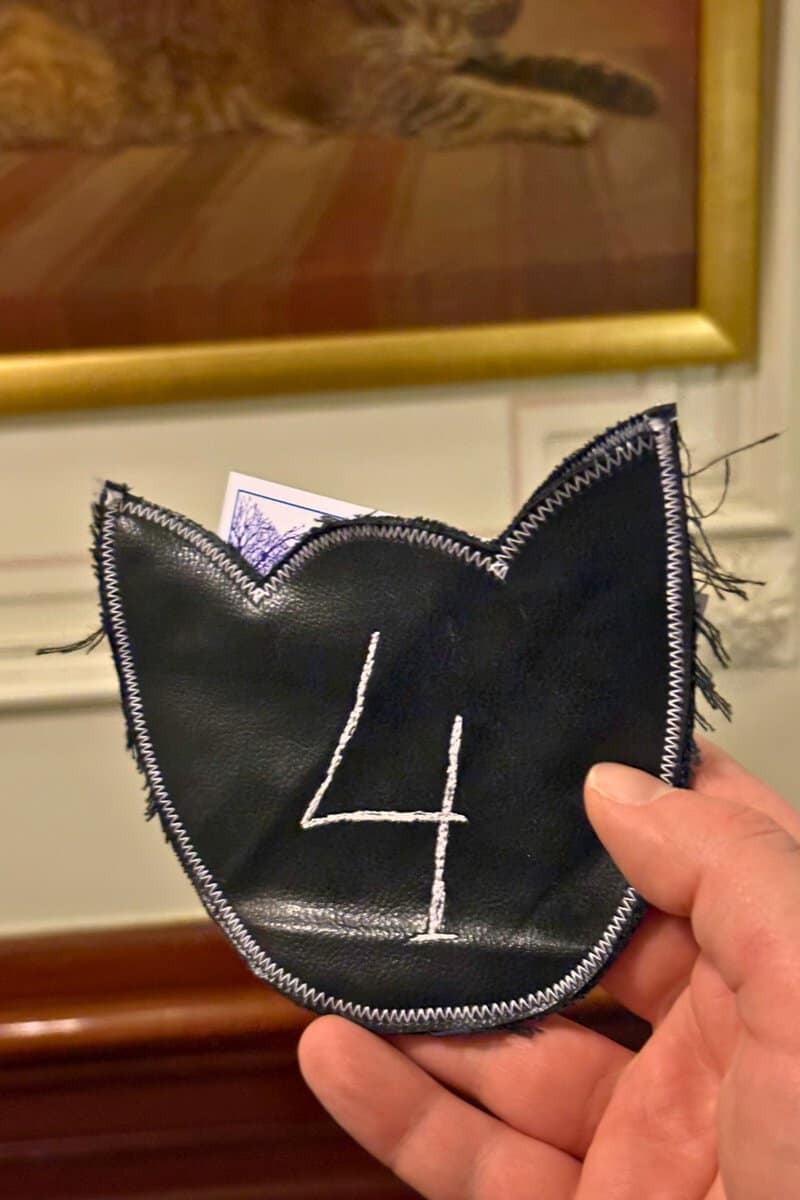 Cat-shaped black leather cloakroom holder marked with a chalk number 4, held up in front of a cat painting at the KattenKabinet. A great place to see cats in Amsterdam.