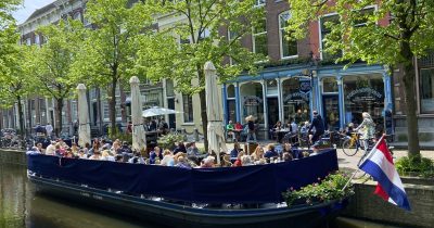 Floating cafe terrace boat with diners and a Dutch flag on a Delft canal.